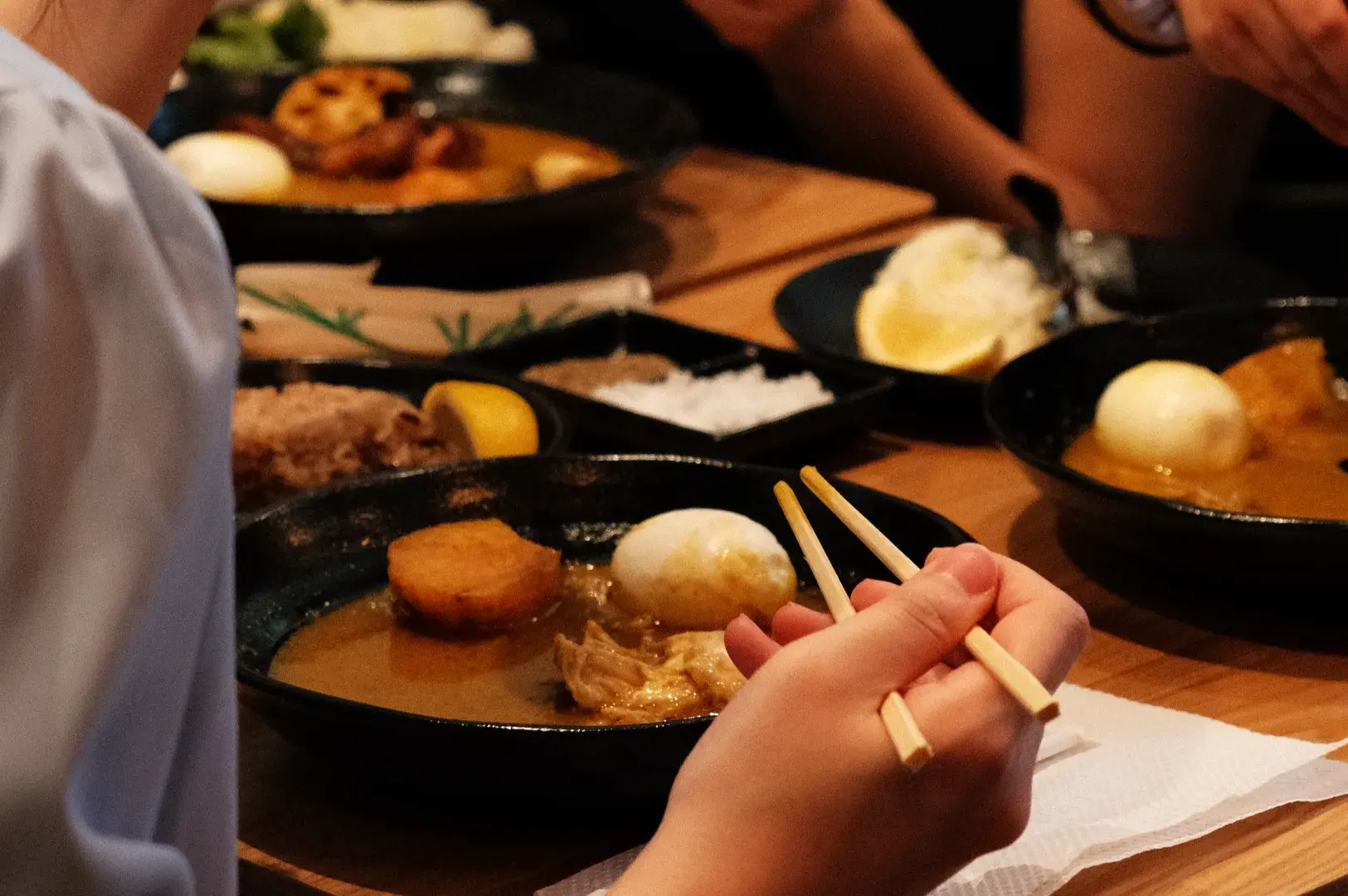 A close-up of hands holding chopsticks above a bowl of food, with various dishes on a table.