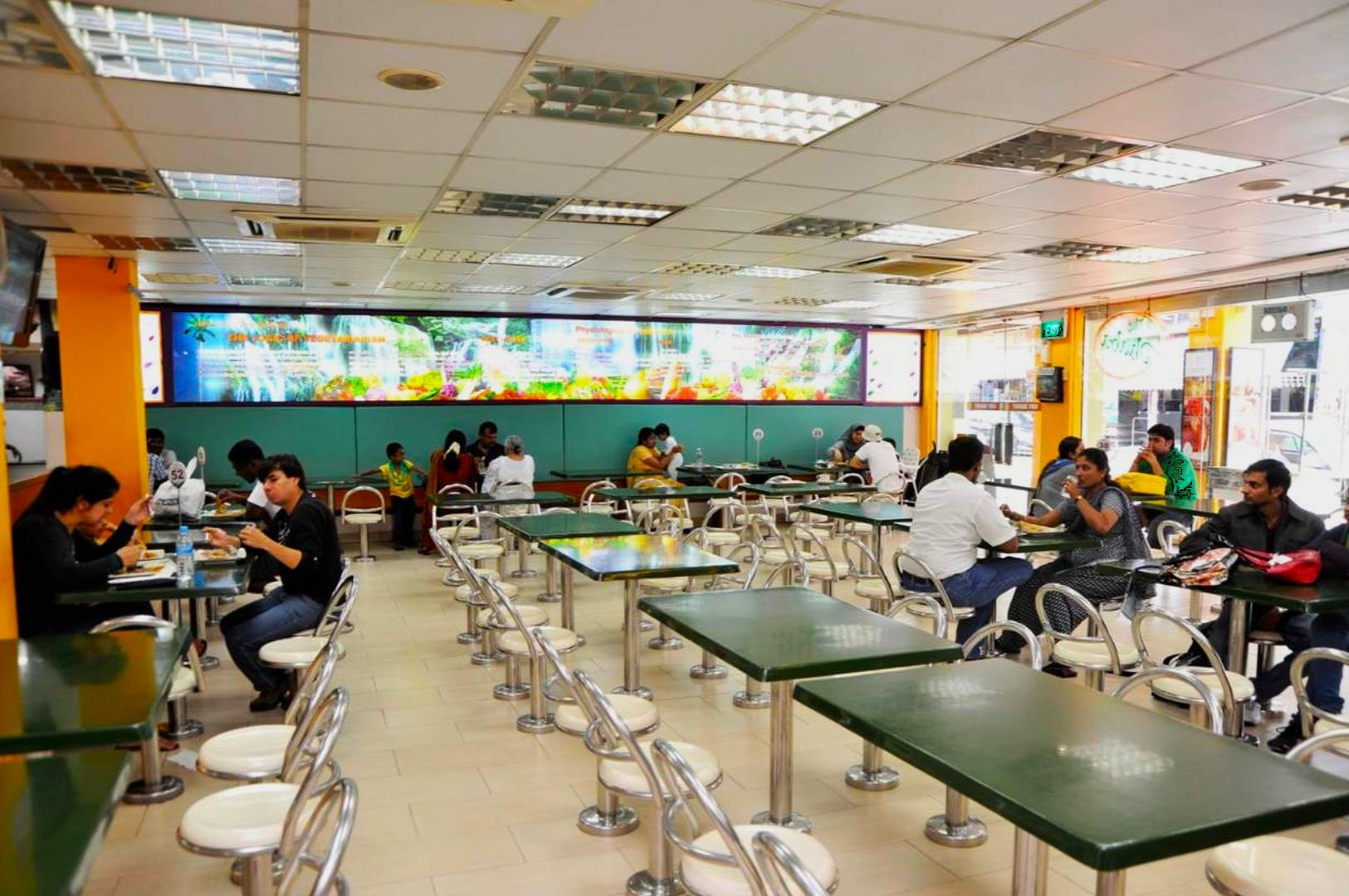 Bright, spacious cafeteria with green tables and white chairs, a colorful mural on the back wall. People dine comfortably, creating a relaxed ambiance.
