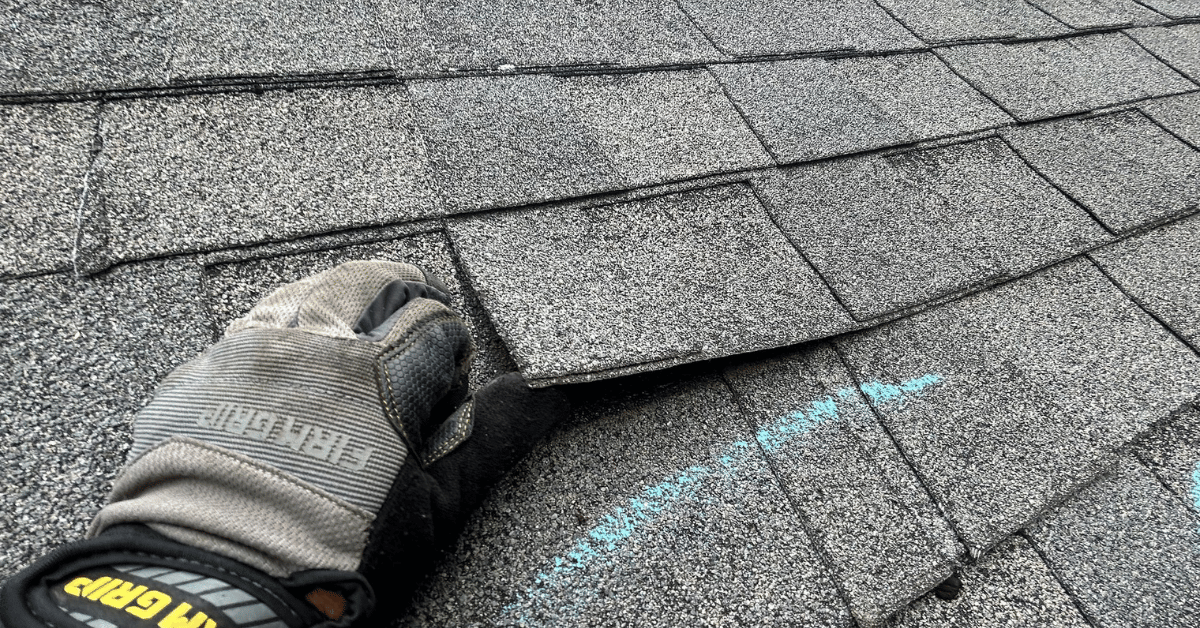Gloved hand lifting a loose asphalt shingle to check for roof damage during an inspection.