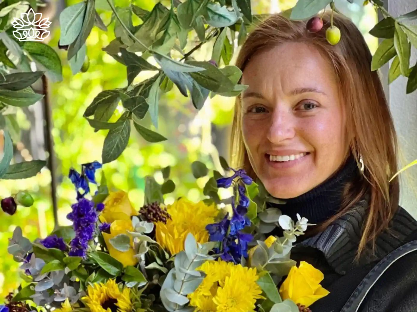 A smiling sa florist holding a vibrant bouquet featuring yellow sunflowers, blue delphiniums, and lush greenery, set against a backdrop of leafy branches. This image represents the New Flower Bouquets Collection, showcasing fresh and beautiful flower arrangements. The collection includes options for deliver flowers with same day delivery. Fabulous Flowers and Gifts.