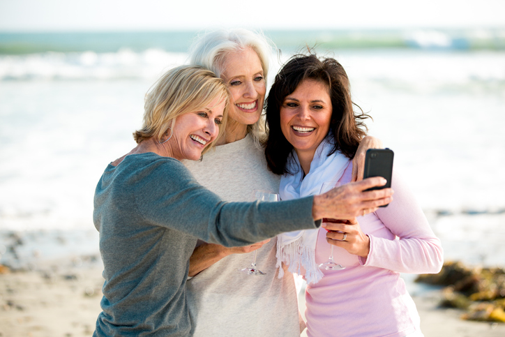 Three mature women smiling on the sand for a selfie.
