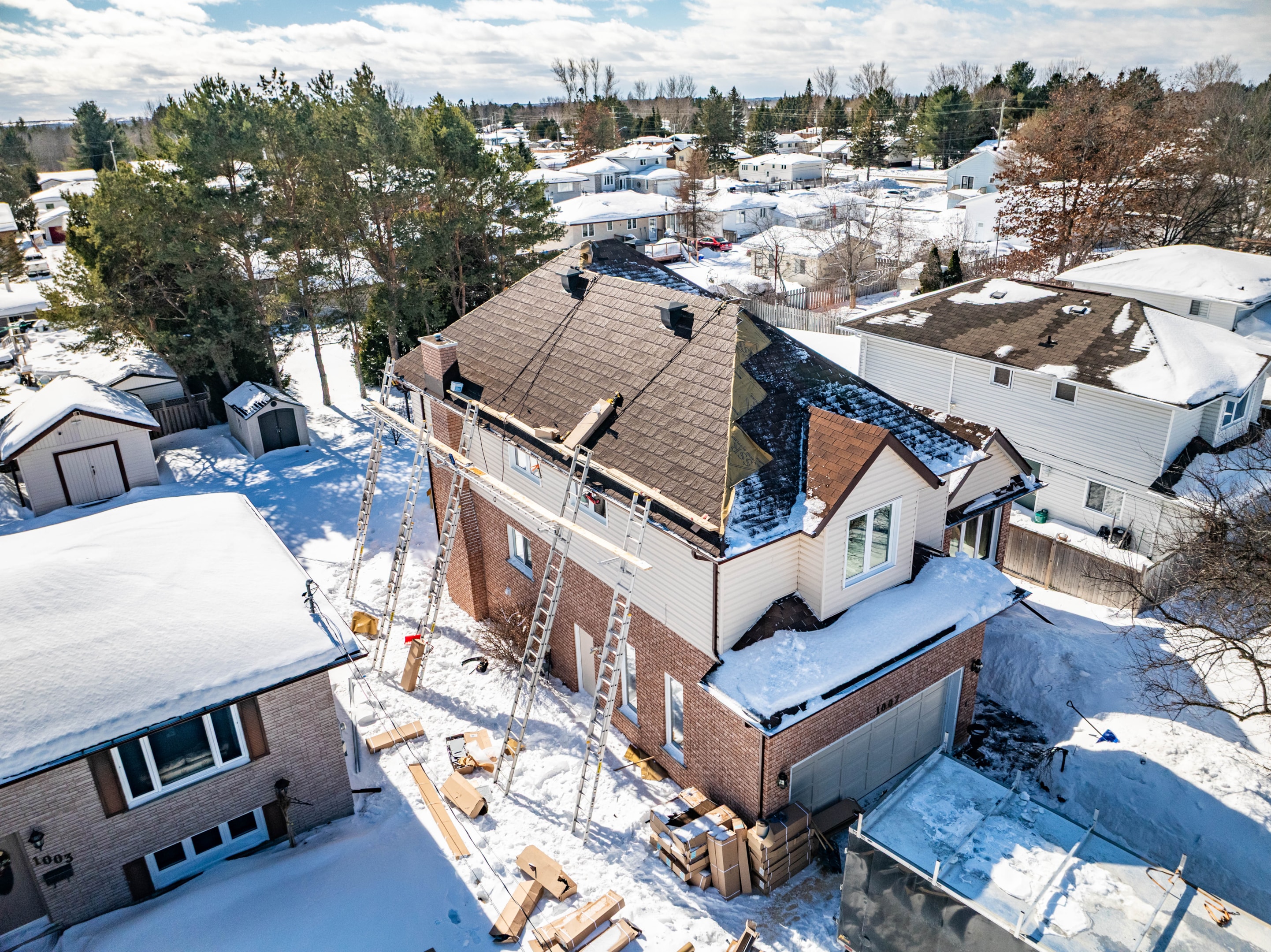 A snow-covered neighbourhood with a house under a metal roof renovation. Ladders and materials are visible, suggesting active construction on a sunny winter day.