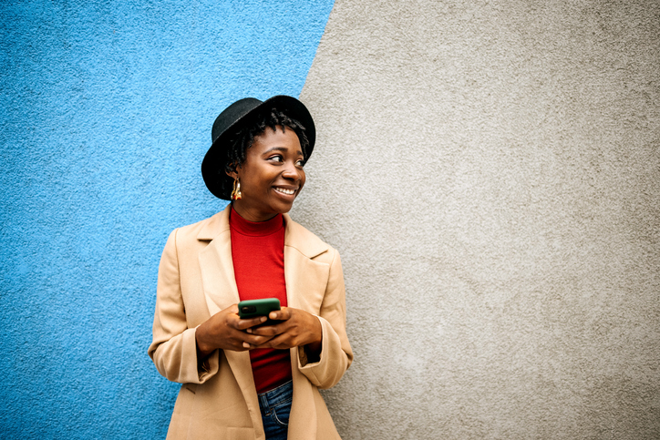 Young woman in a black hat smiling against a blue and grey wall.