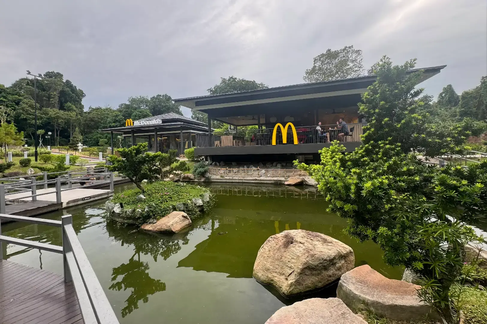 An angled view across the pond showing large boulders along the water's edge, lush green trees framing the right foreground, and diners seated on the covered outdoor balcony of the pavilion adorned with the yellow McDonald's logo.