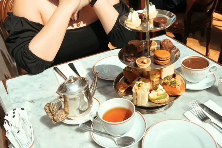 A three-tiered silver stand displays an assortment of afternoon tea delicacies, including macarons, tartlets, and savory sandwiches on a marble tabletop. Beside it, a silver teapot and cups of tea and hot chocolate complete the elegant spread.
