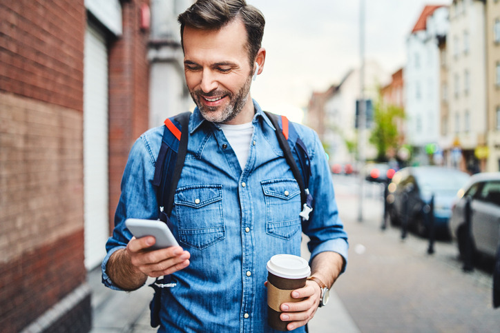 Dark-haired man carrying a backpack smiling at his cell phone.