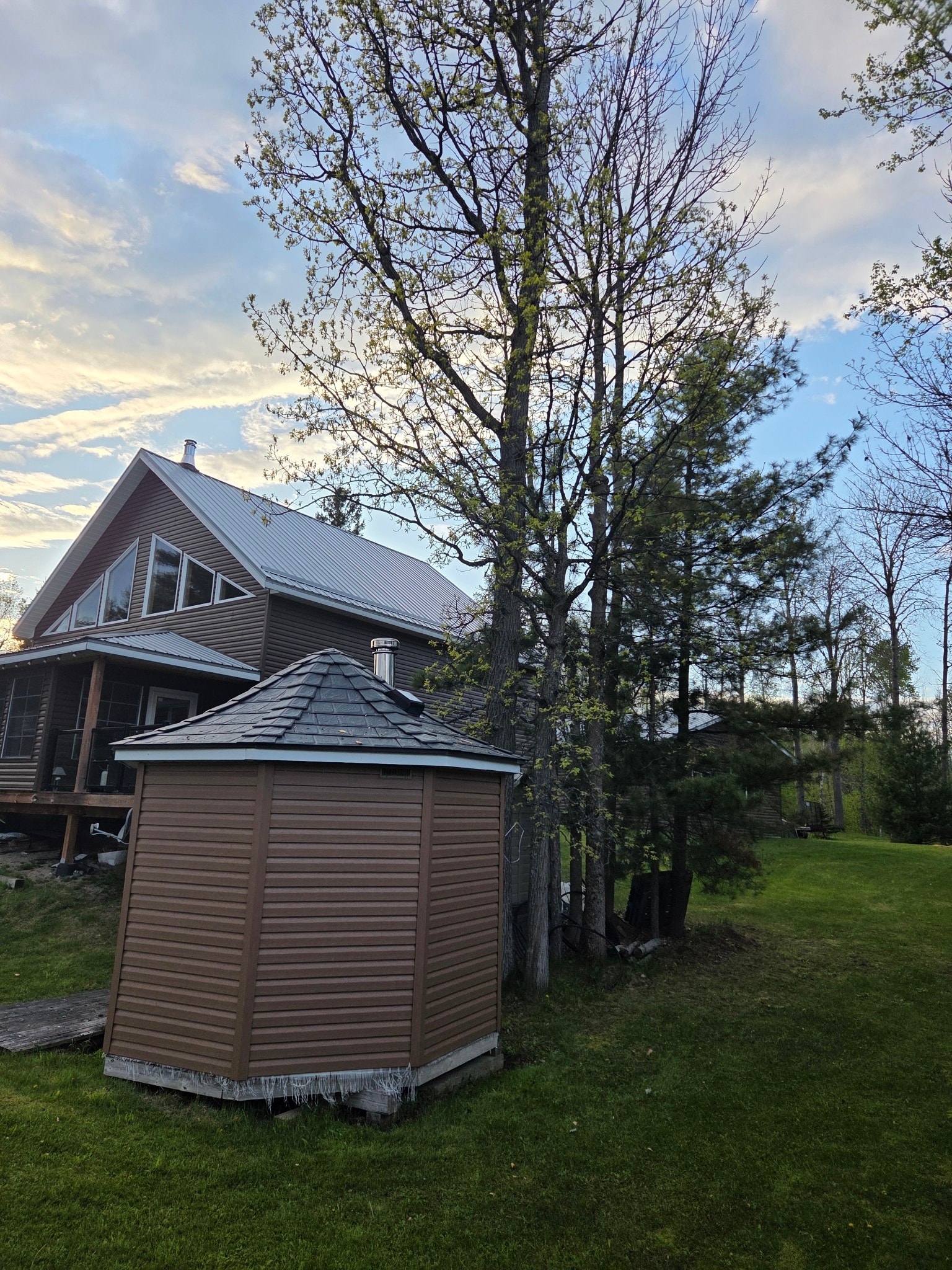A residential house featuring a shed in the yard with sloped metal roofing, set against a backdrop of greenery and open space.