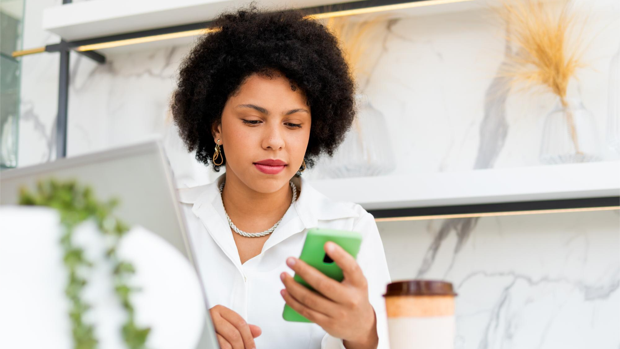 A woman with curly hair and a white blouse looks at her green smartphone while working at a laptop in a modern cafe. This image reflects how mobile communication and Business SMS allow for on-the-go productivity using business messaging tools.