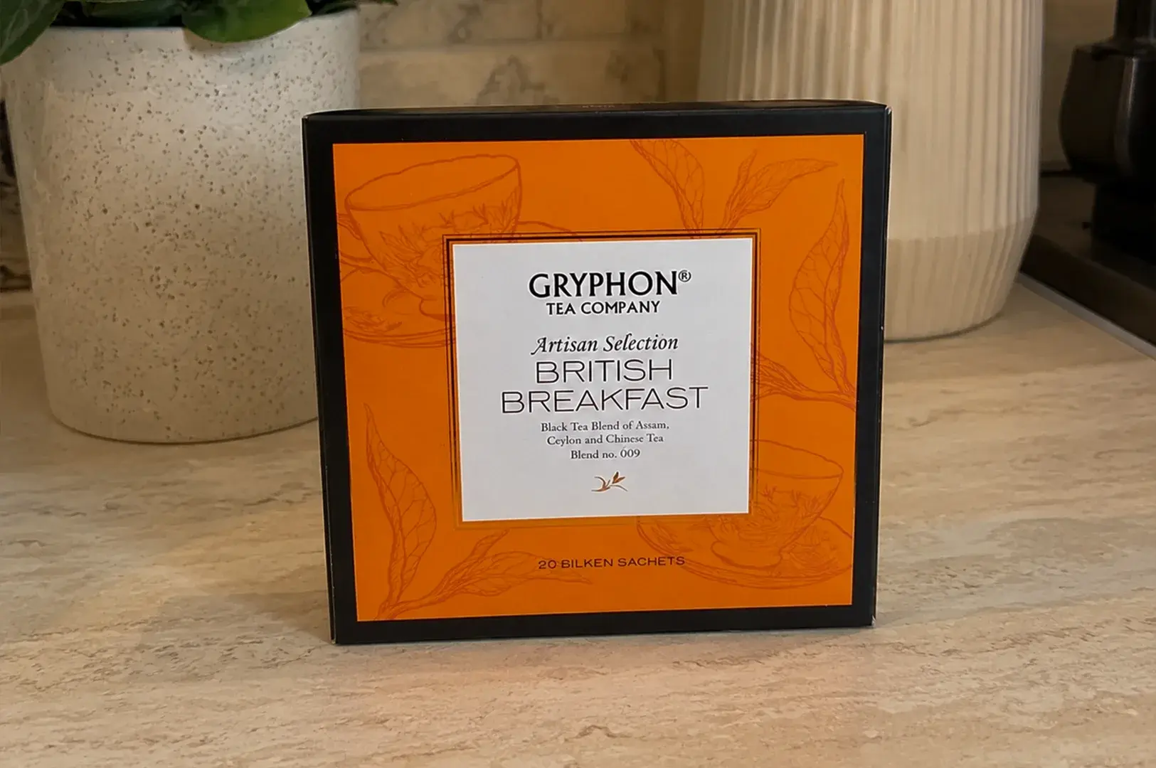 A square tea box of Gryphon Tea Company's Artisan Selection "British Breakfast" blend sits prominently on a beige stone-textured countertop. The front panel of the box features a bold, bright orange background adorned with subtle line art drawings of teacups and tea leaves, framing a clean white central label that describes the tea as a black tea blend of Assam, Ceylon, and Chinese tea. The box is flanked by a speckled off-white ceramic planter containing a green houseplant on the left, and a cream-colored ribbed ceramic vase in the background.