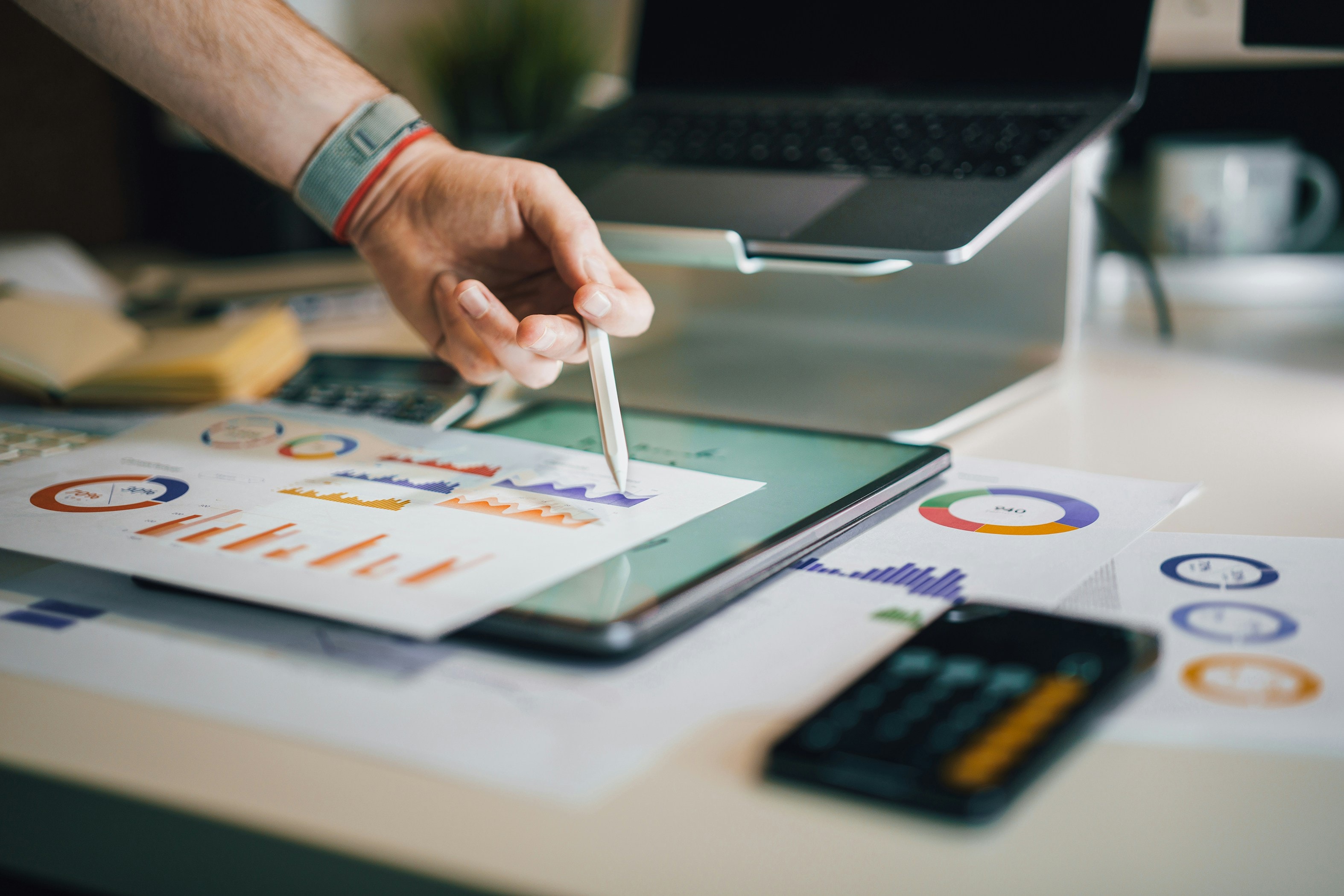 Hand with a pen showing something on paper with pie charts and a tablet, calculator and a laptop around to it