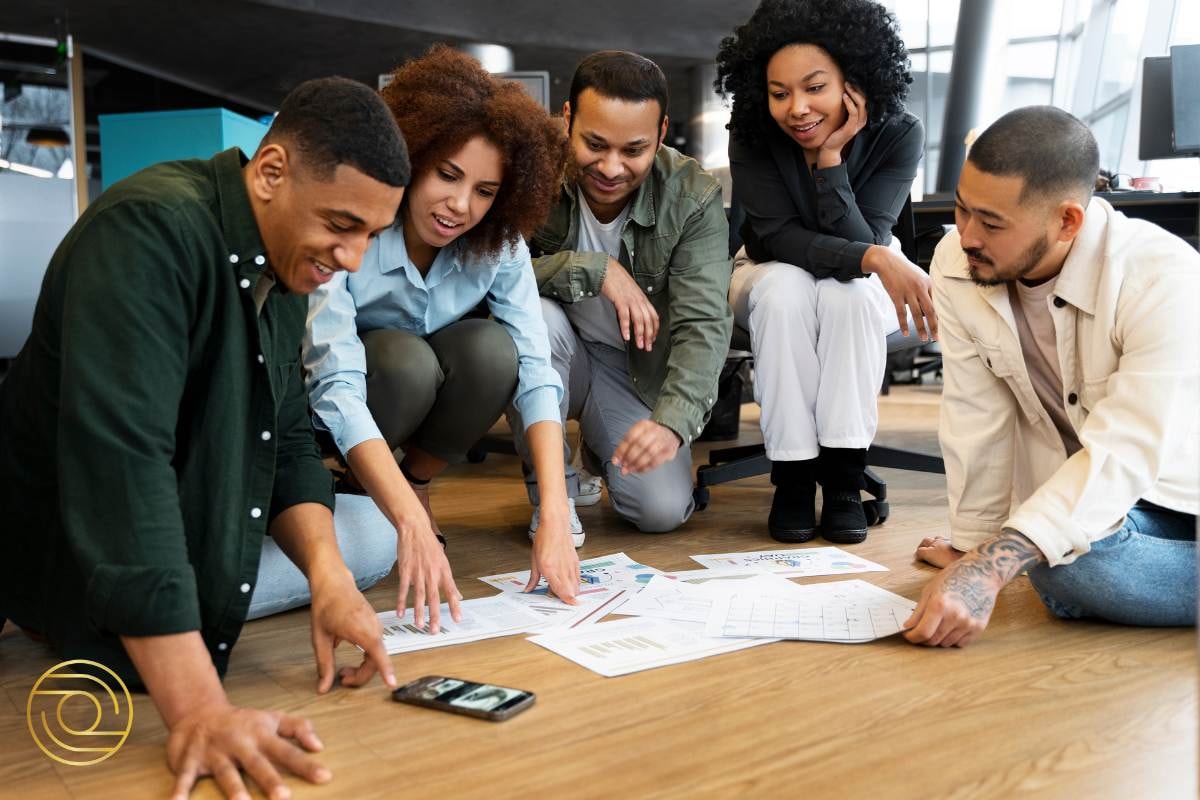 Collaborative team effort as members gather around papers on the floor, discussing ideas and using a phone for reference.