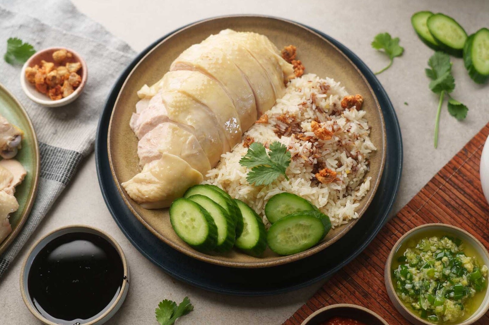 Sliced poached chicken and fragrant rice garnished with crispy shallots and cilantro, accompanied by cucumber slices, soy sauce, and green chili sauce.