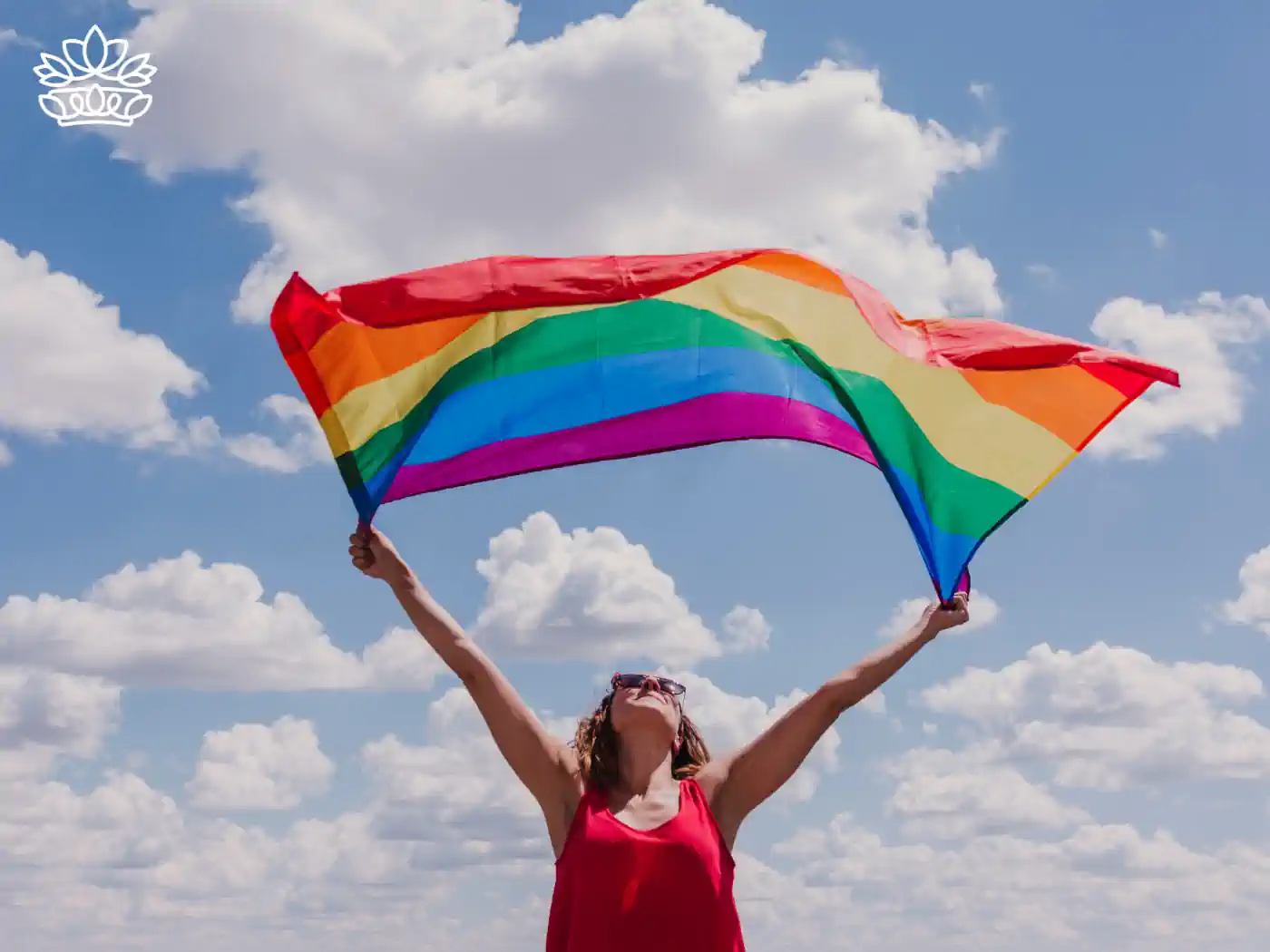 A person waving a rainbow flag under a bright blue sky, symbolizing pride and celebration. Fabulous Flowers and Gifts - Pride Collection.