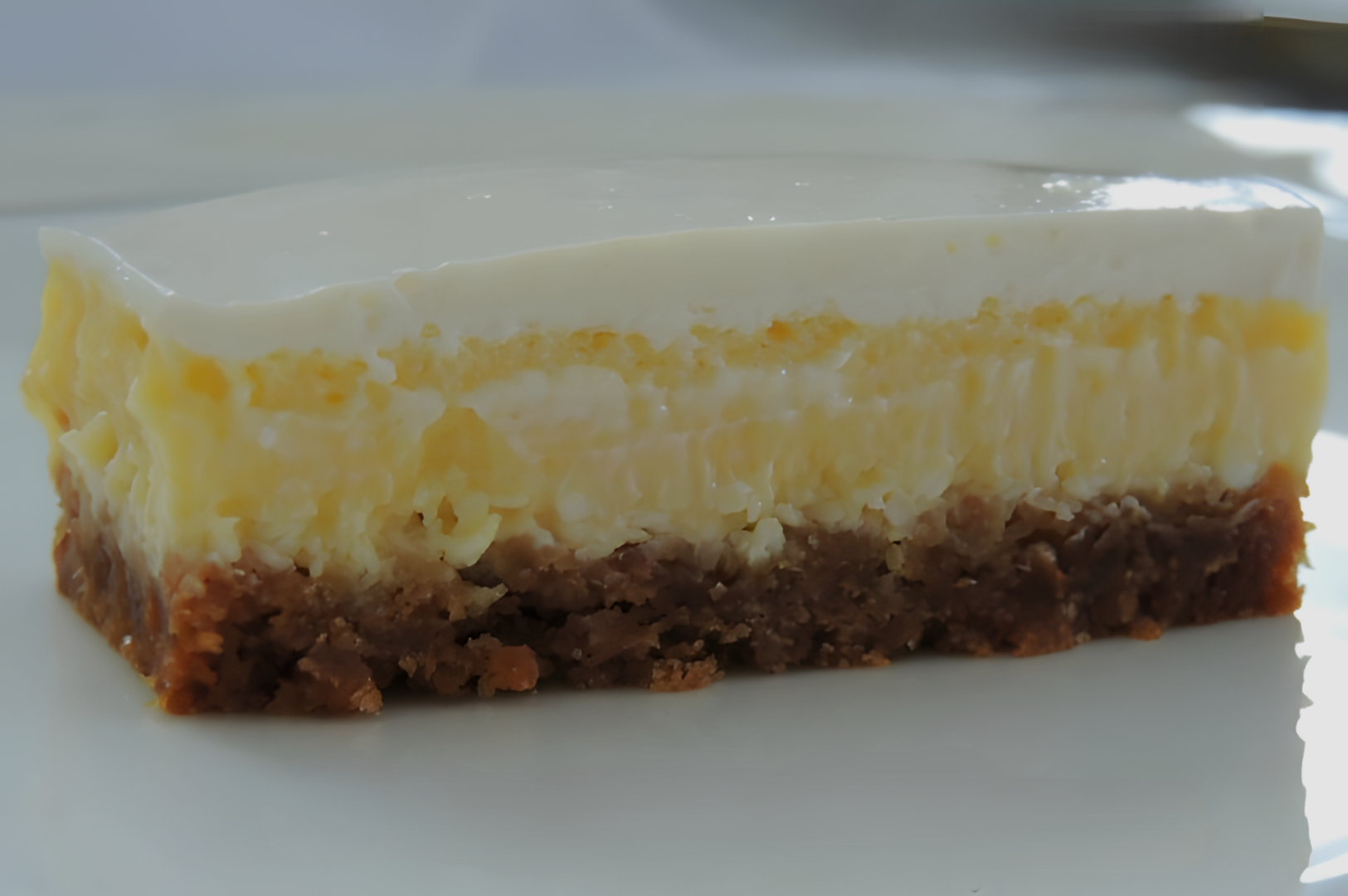 A creamy cheesecake slice on a white plate, featuring a brown crumbly crust, a yellowish cheese layer, and a smooth white cream topping.