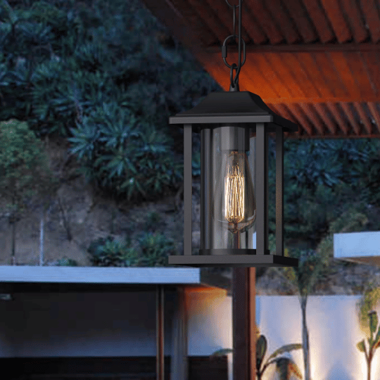 Black outdoor hanging lantern with clear glass panels and a glowing filament bulb, suspended under a wooden pergola at dusk.