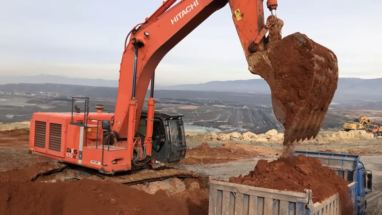 An orange Hitachi excavator scooping up a large amount of dirt and unloading it into the back of a blue dump truck, with a mountainous landscape and mining operations in the background