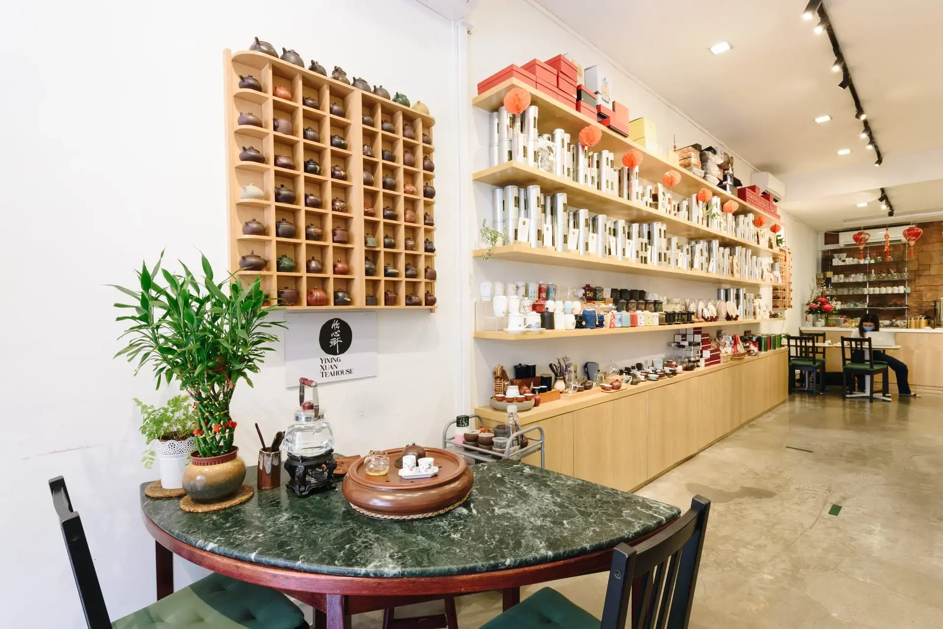 This image showcases the interior of Yixing Xuan Teahouse, featuring an organized wall display of clay teapots and shelves lined with tea canisters. In the foreground, a round marble table is set with a traditional tea brewing station, complete with a glass kettle and small tasting cups.