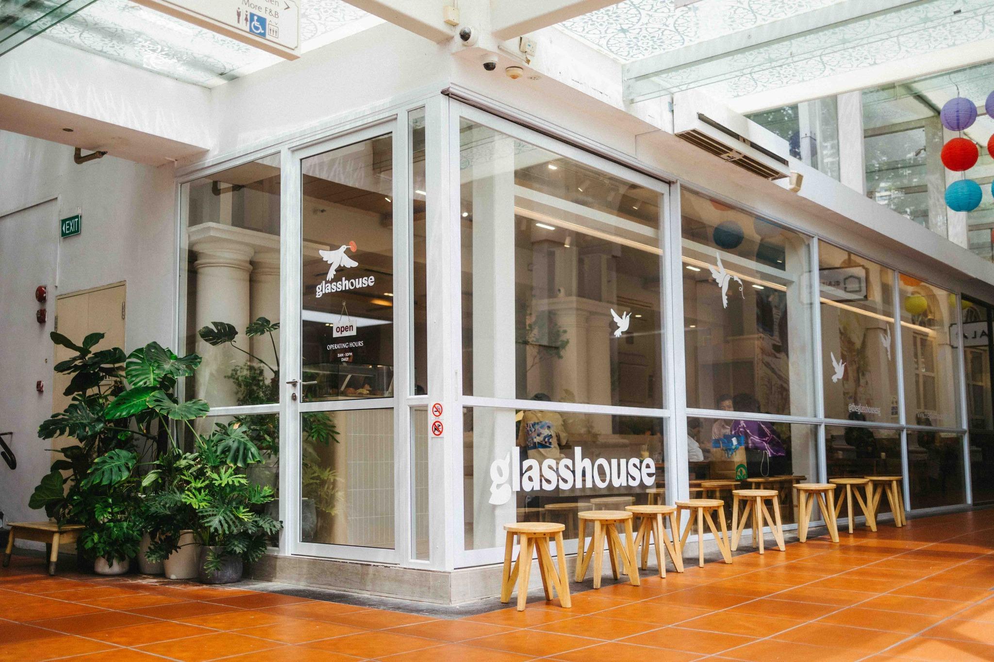 A modern cafe with large glass walls and white framing stands at a corner, accented by lush potted greenery. A row of simple wooden stools lines the exterior under a bright, skylit walkway with orange tiled flooring.
