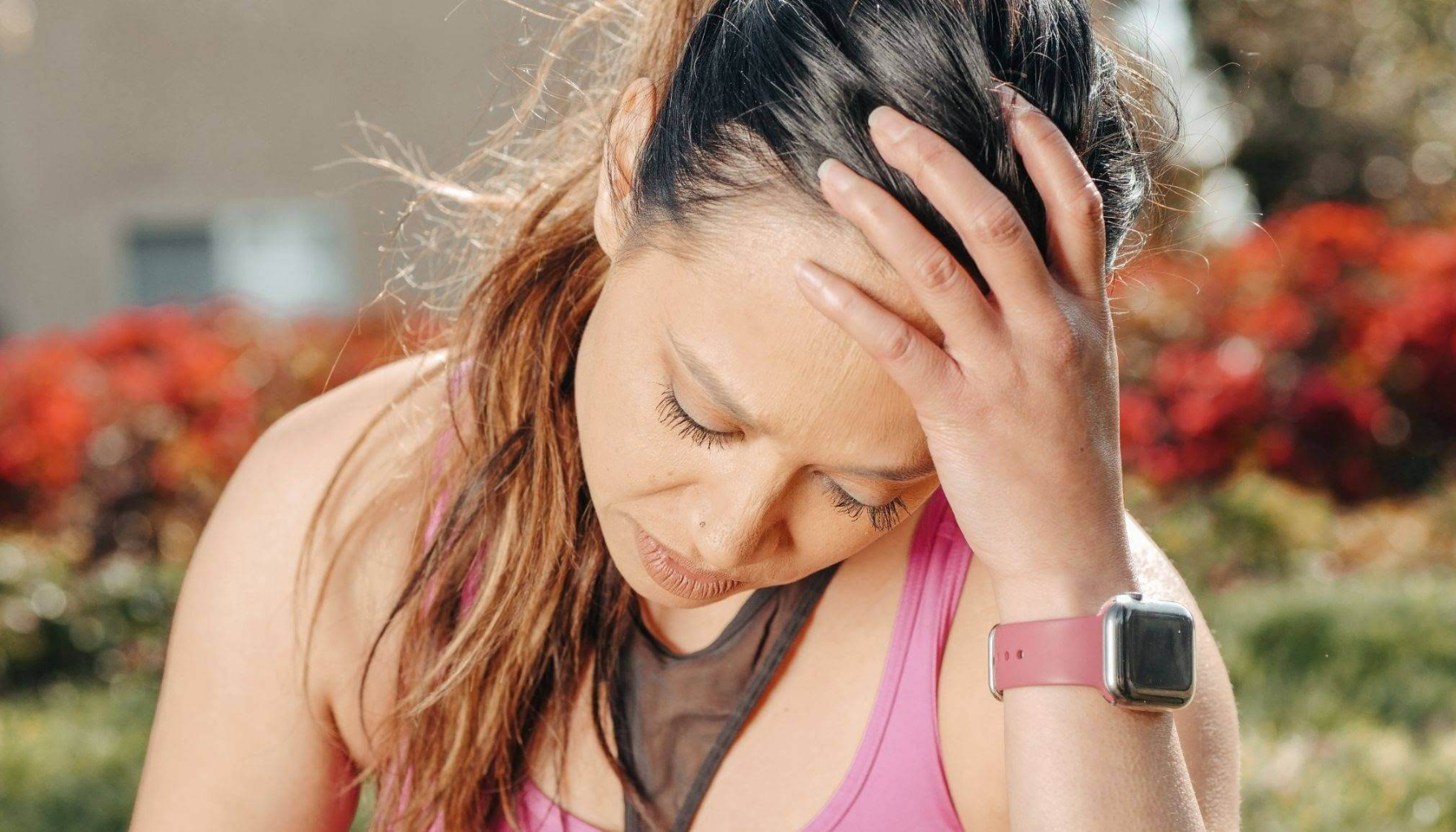 "Kvinne i treningsklær holder hodet med hånden, tydelig plaget av hodepine eller utmattelse utendørs."