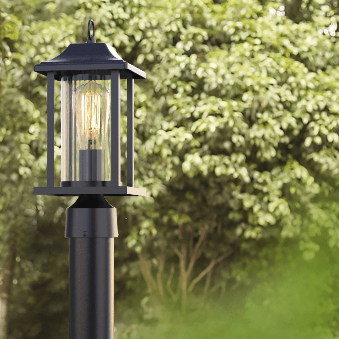 Black outdoor post lantern with clear glass panels and glowing filament bulb, set against a background of green trees.