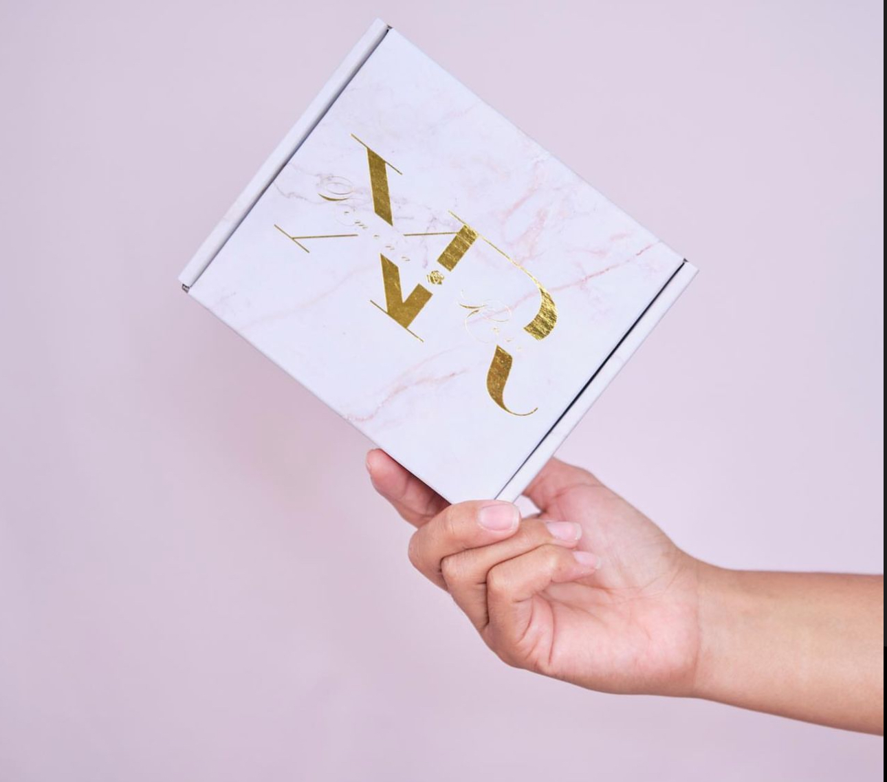 A pink background featuring a hand holding a pink packaging box with 'XR' for Ximena Rose.