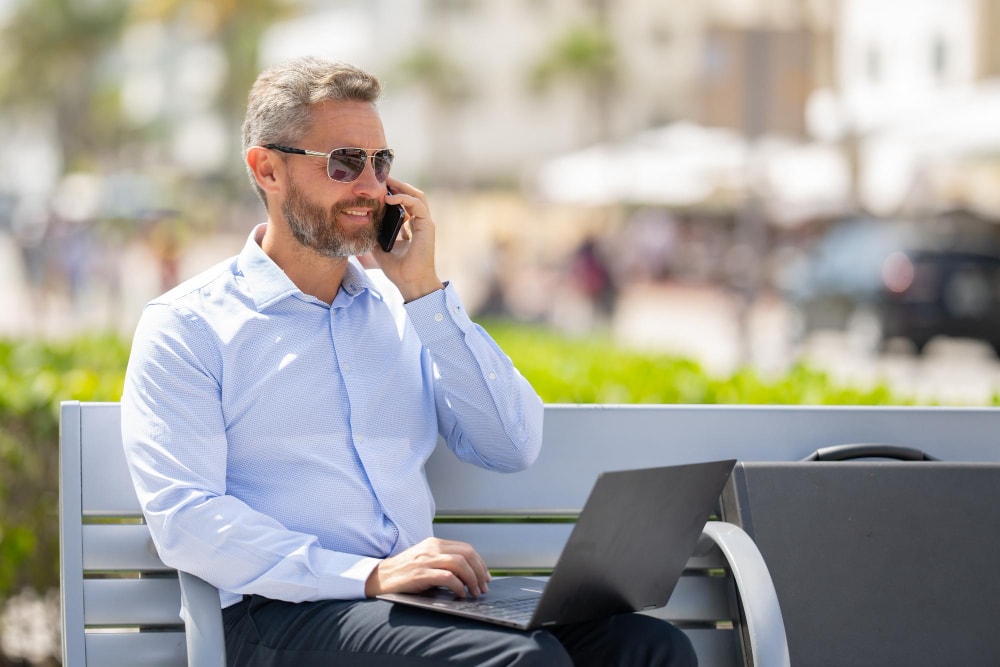 Businessman sitting on a bench with a laptop and taking phone calls in the park.