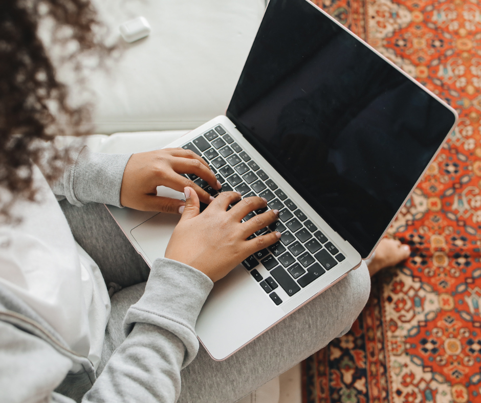 a woman wearing sweats sitting on her couch typing on a laptop