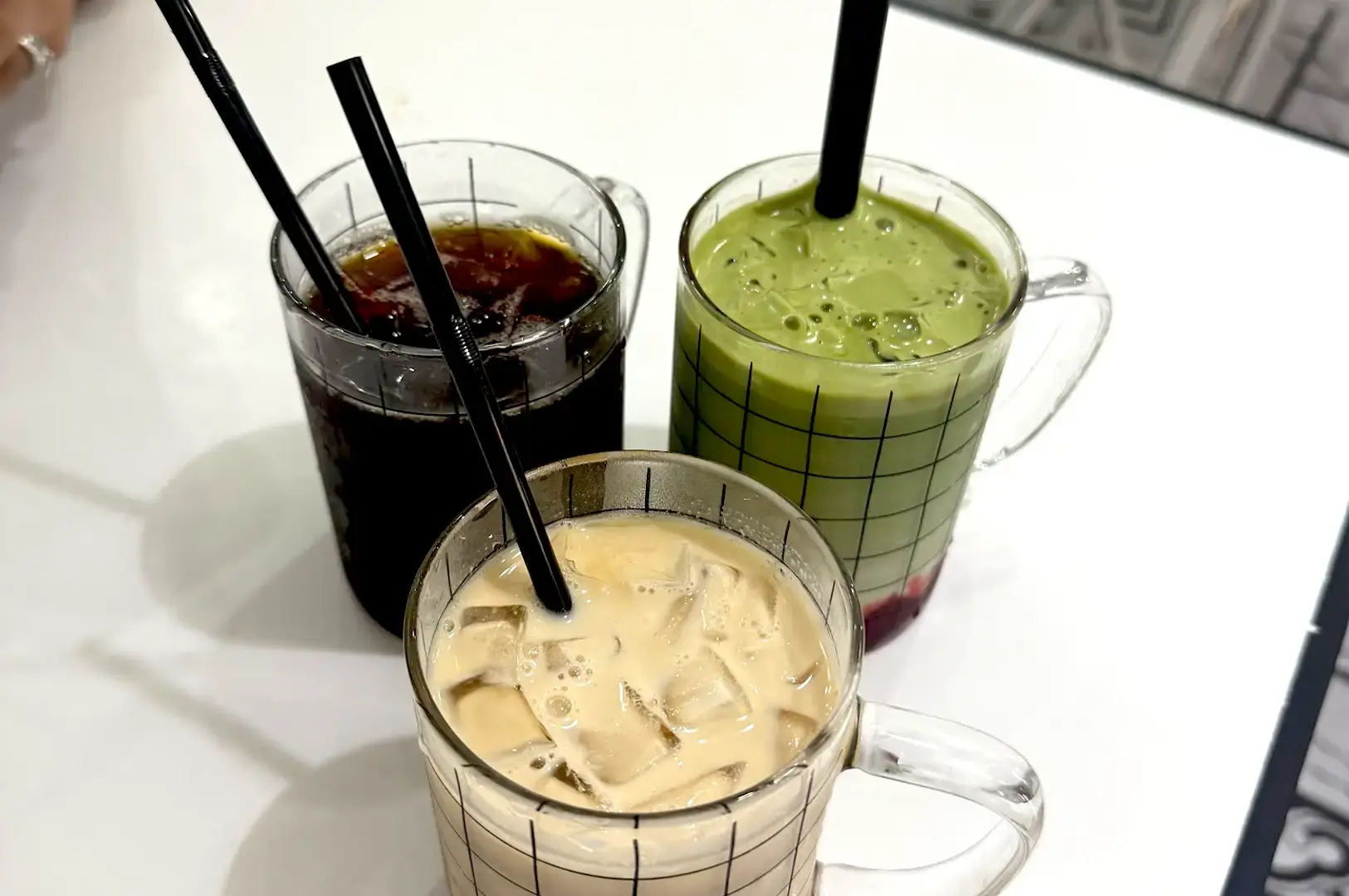 Three clear glass mugs featuring a grid pattern and filled with iced coffee, iced milk tea, and a layered matcha drink with black straws at Cafe Monochrome.