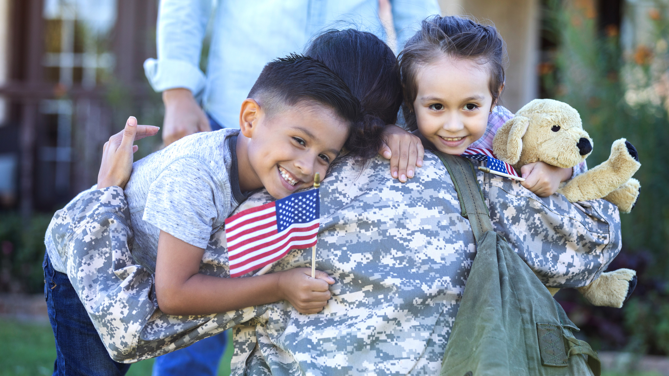 Military mom hugging her children while they smile and hold American flags and stuffed animals.