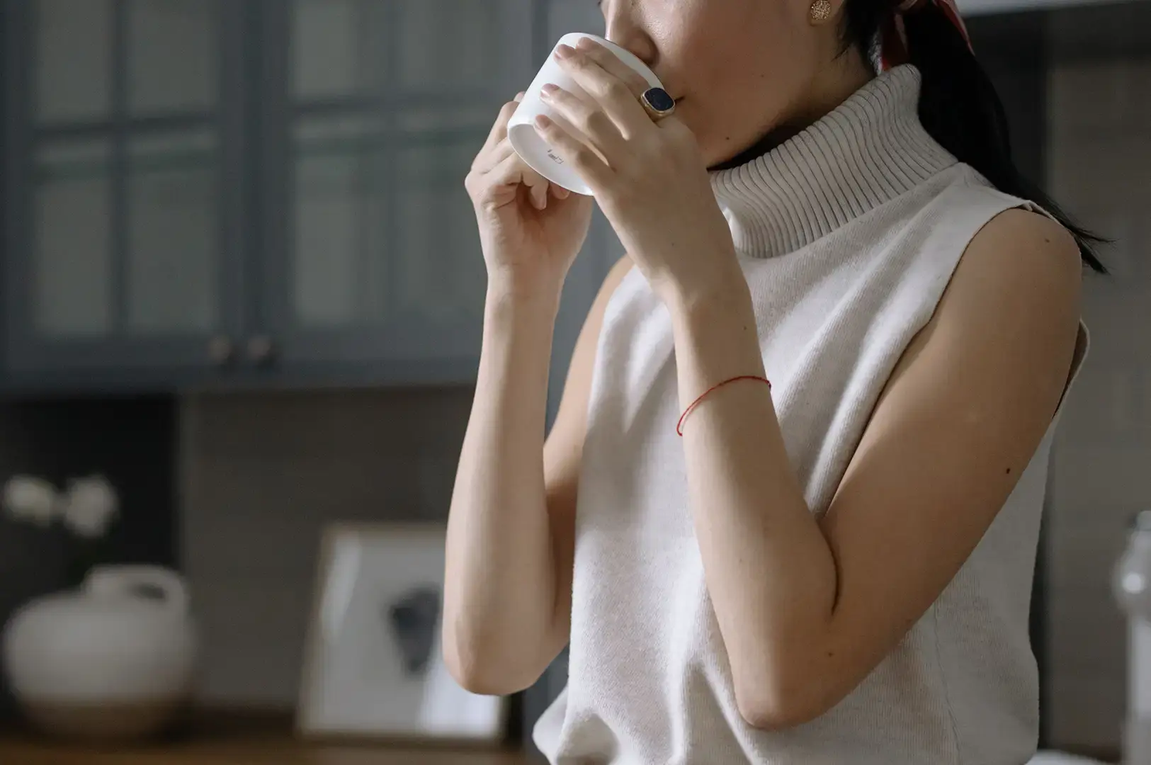 A close-up, cropped view of a woman in a kitchen setting taking a sip from a white ceramic mug held with both hands. She is wearing a sleeveless white knit turtleneck sweater, a thin red string bracelet on her left wrist, and a large ring with a dark blue stone on her right hand. The background features muted blue kitchen cabinetry and a soft focus white ceramic vase, creating a calm and mindful atmosphere.