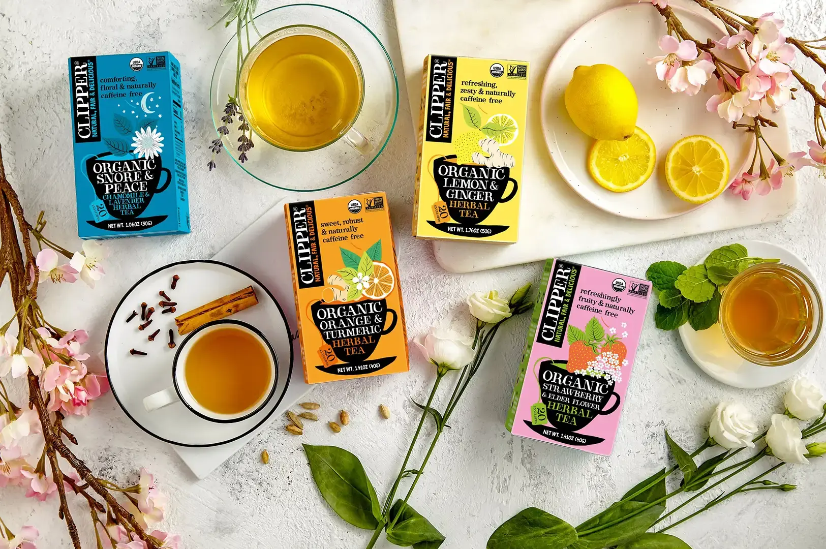 An elegant top-down flat lay featuring four vibrant Clipper Herbal Tea boxes—"Snore & Peace", "Lemon & Ginger", "Orange & Turmeric", and "Strawberry & Elderflower"—arranged on white marble cutting boards and surrounded by fresh white flowers, pink cherry blossoms, loose spices, lemon slices, and two warm cups of brewed tea.