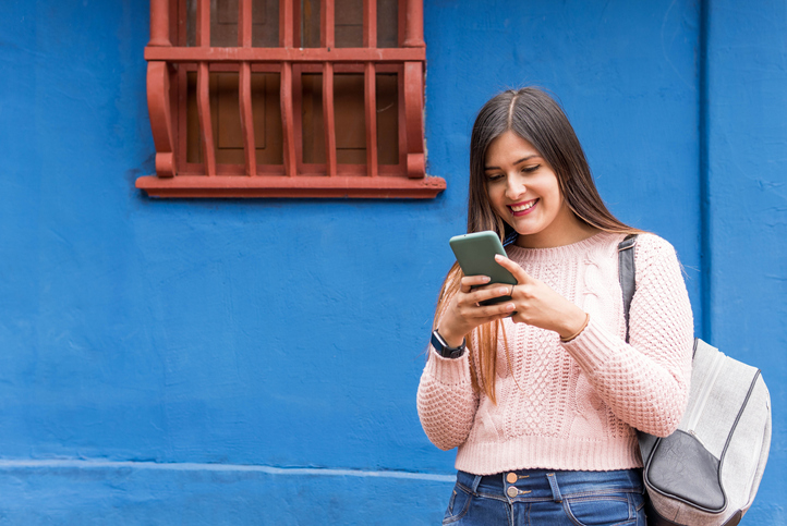 Cheerful young woman in a pink sweater standing next to a blue wall and checking her cell.
