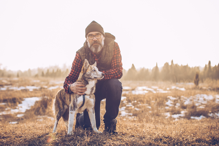 Man wearing a cap and a flannel shirt petting his dog in a field. 