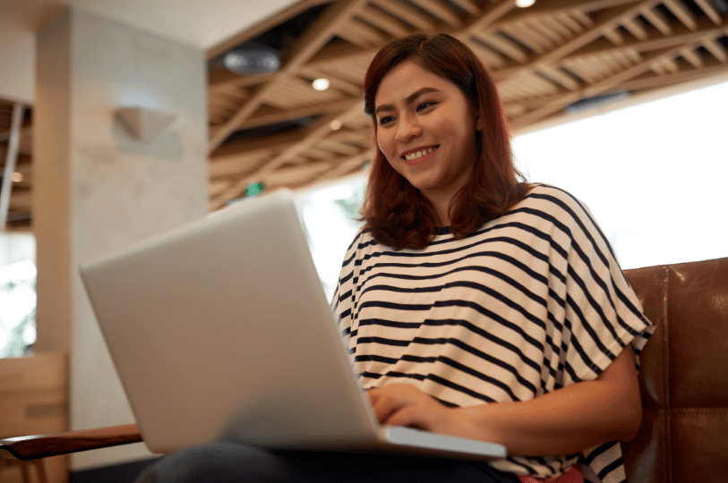 A woman smiling while looking at her laptop in a cafe setting, representing a customer successfully finding and booking a table at a restaurant online.