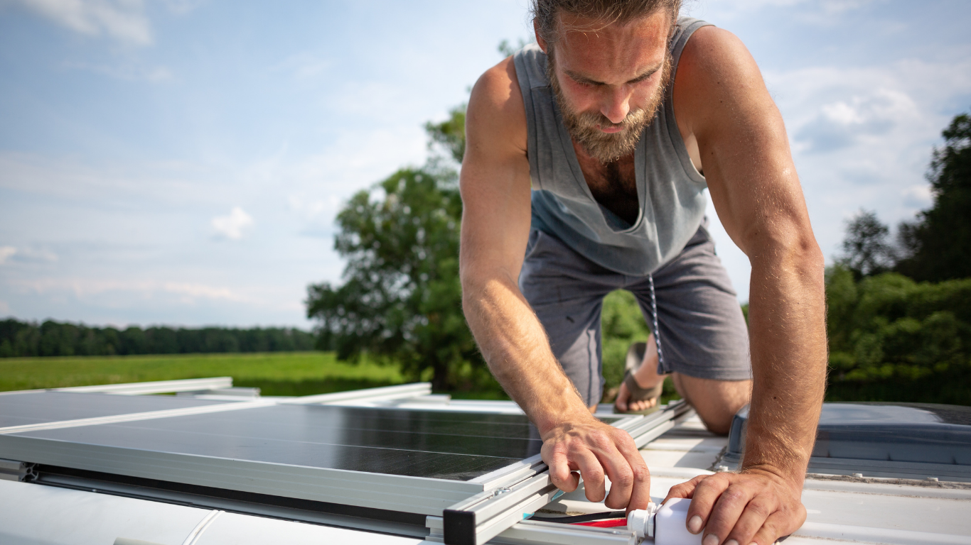 Mann montiert ein Solarpanel auf dem Dach eines Campers an einem sonnigen Tag in ländlicher Umgebung
