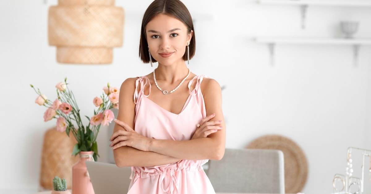 A woman in a light pink dress stands confidently with her arms crossed, symbolizing the professionalism and approachability of a certified tax planner.