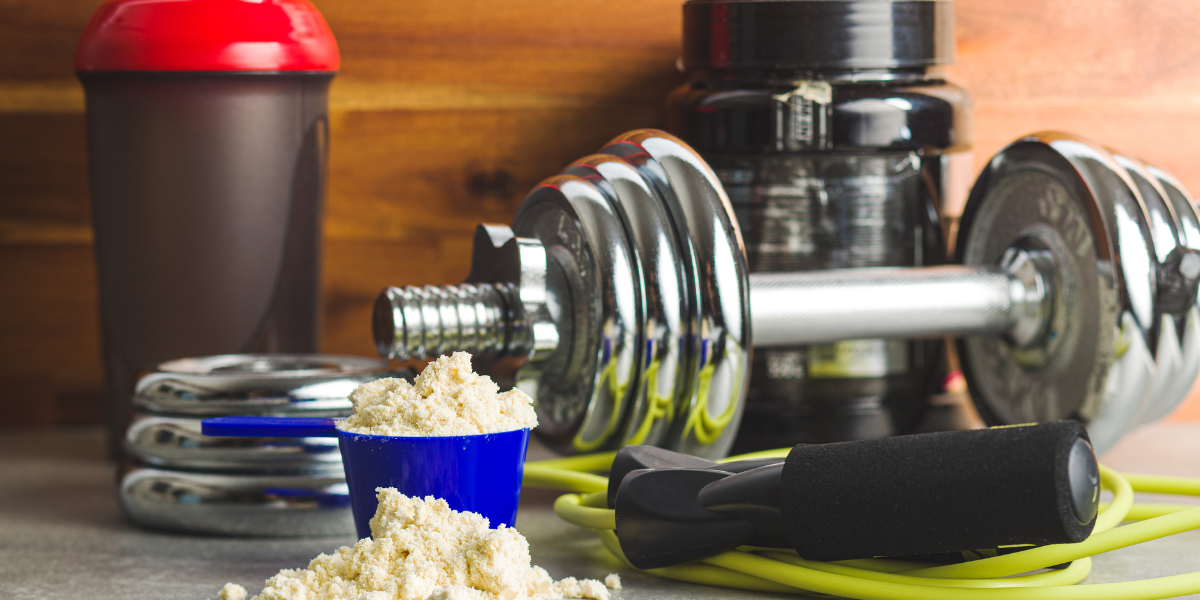 Drink protein powder to reach your daly protein targets. Image of protein powder overflowing from a protein scoop next to a dumbbell, jump rope, protein shaker, and container of protein powder.