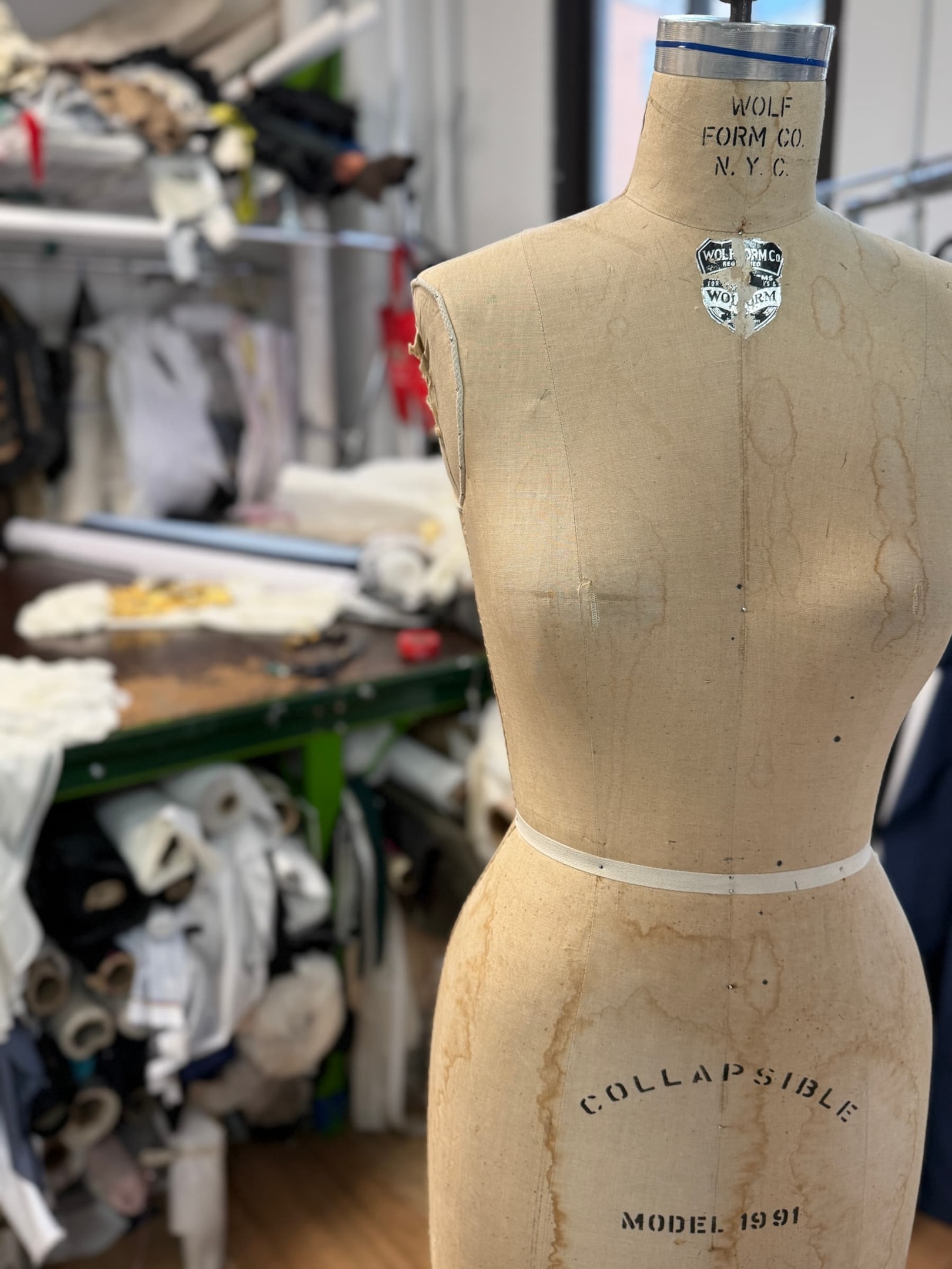 Sewing dress form in front of a cutting table of a dressmaker shop with clothing, fabric, crafts and other items in the background