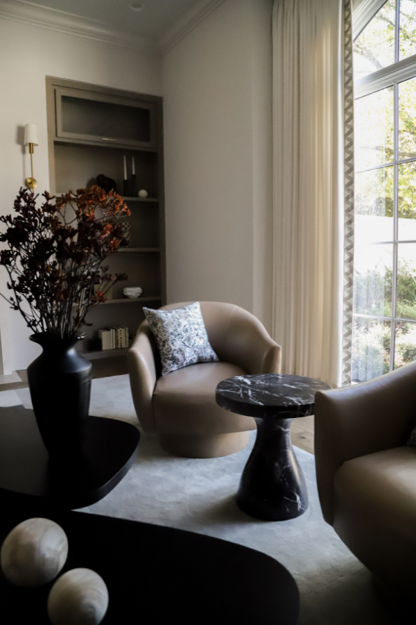 A neutral living room featuring curved leather chairs, a black marble side table, soft floor-to-ceiling drapery, and a minimalist built-in shelf.