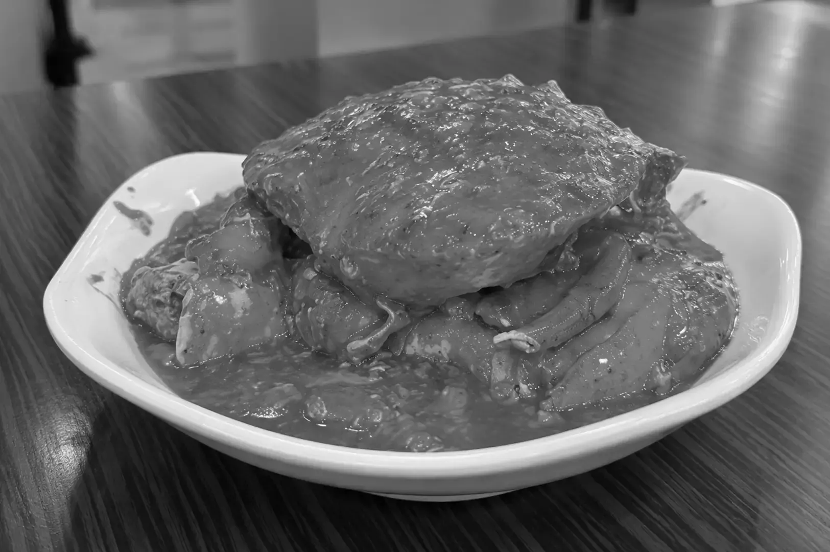 A whole crab covered in thick, glossy sauce and arranged with its legs and claws on a white oval plate, photographed closely on a dark wooden table.