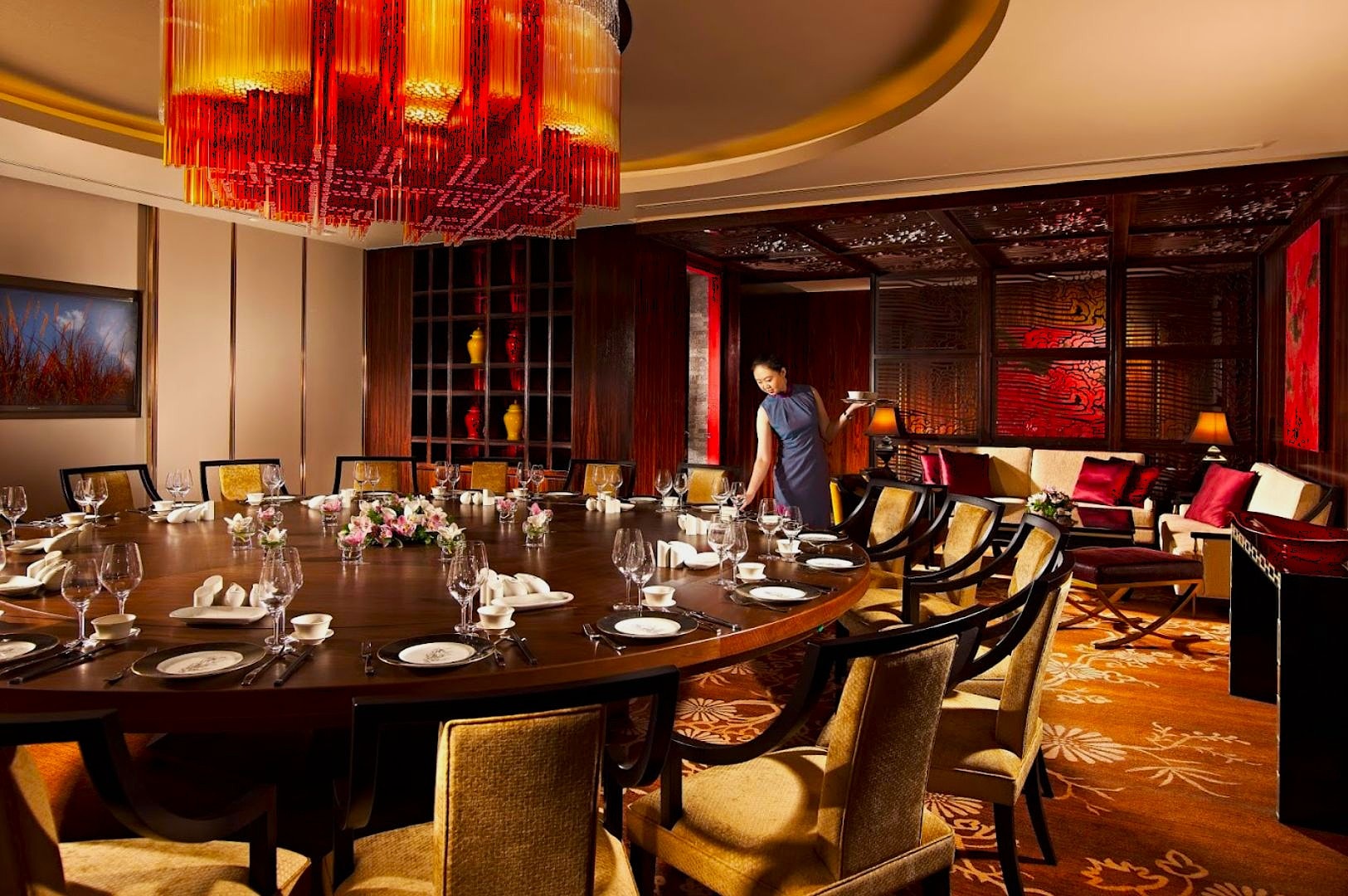 Luxurious dining room with a large, round wooden table set for a banquet. Elegant red chandelier overhead, a staff member arranging chairs, cozy ambiance.