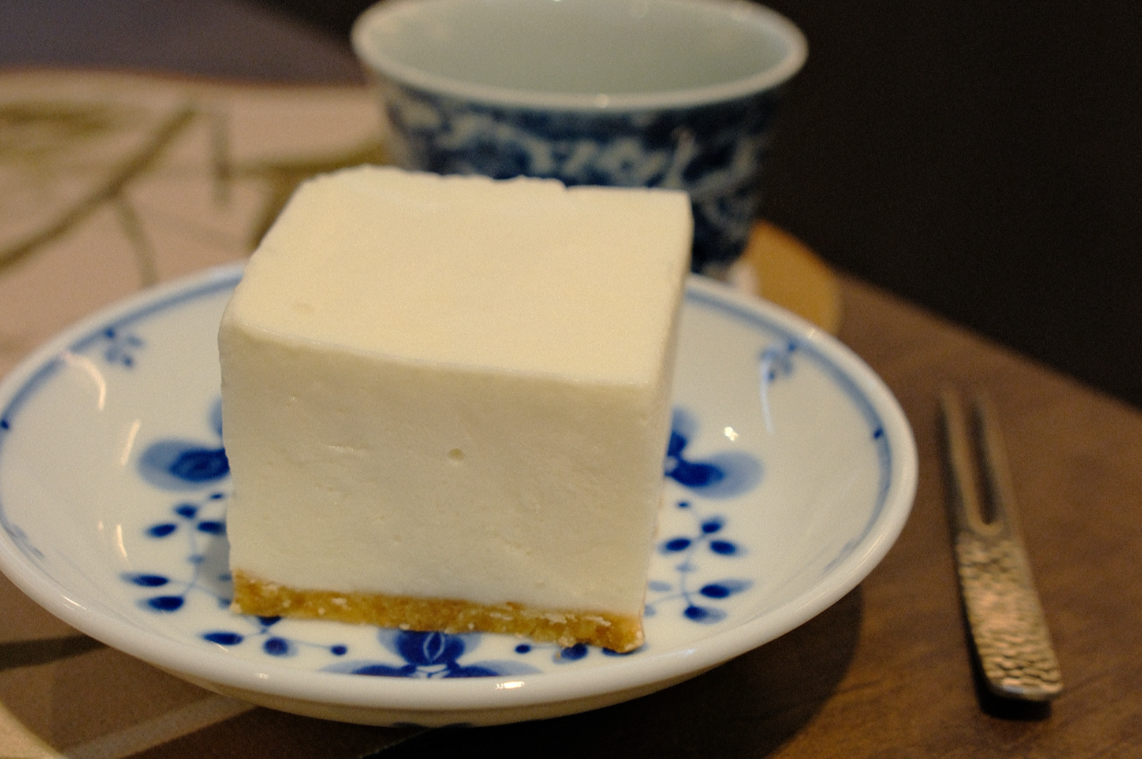 A slice of creamy cheesecake sits on a white and blue floral plate, next to a textured fork. A detailed ceramic cup is in the background.