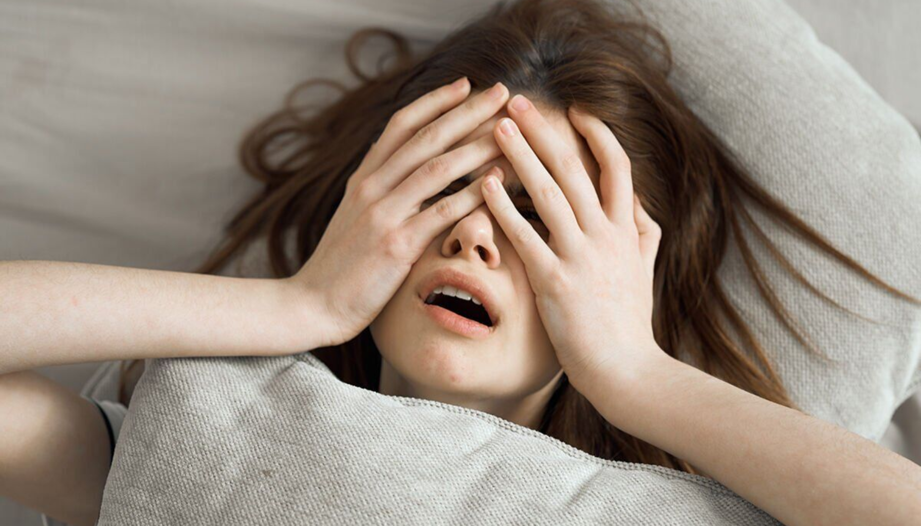 Kvinne ligger i sengen med hendene foran ansiktet og åpen munn, ser utmattet og stresset ut.