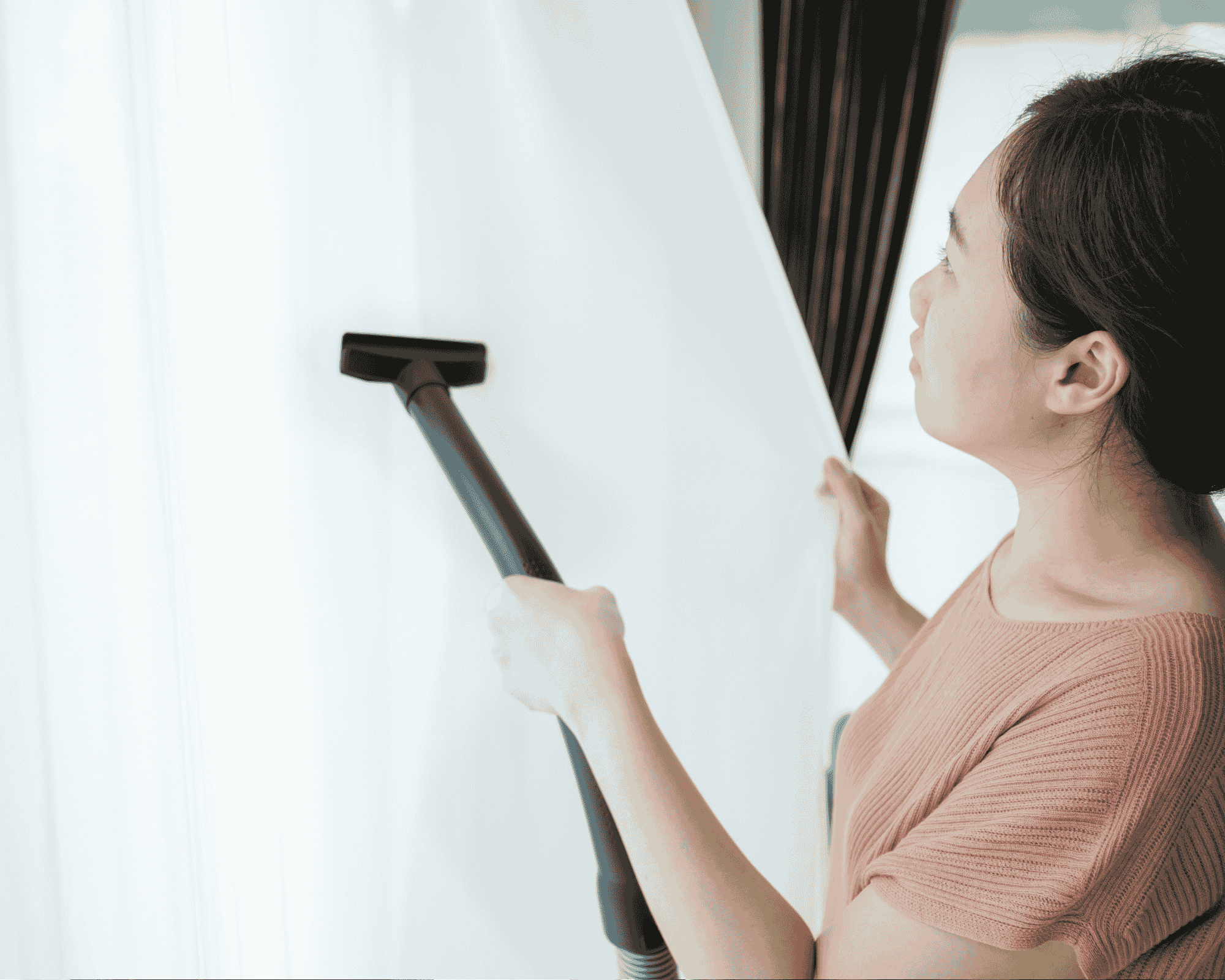  Professional curtain cleaning service in action – a woman carefully vacuuming sheer white curtains at home using a modern upholstery attachment. This image highlights deep curtain cleaning techniques used to remove dust, allergens, and pollutants, ideal for allergy prevention and maintaining fresh indoor air quality. Suitable for residential and commercial curtain maintenance solutions. 