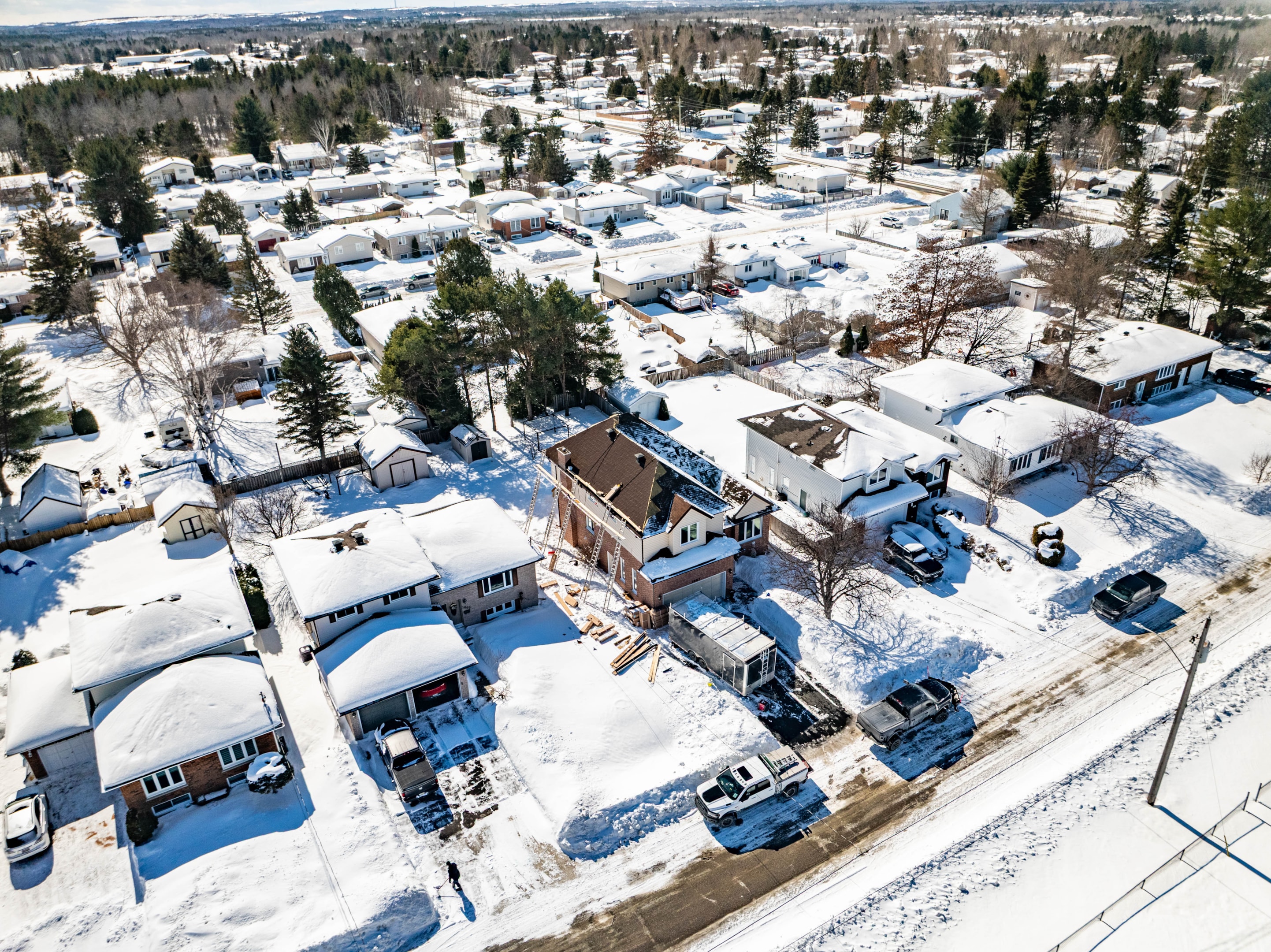 An aerial view of a snowy suburban neighbourhood with houses, snow-covered metal roofs and yards. A construction site is visible in the center, surrounded by trees.