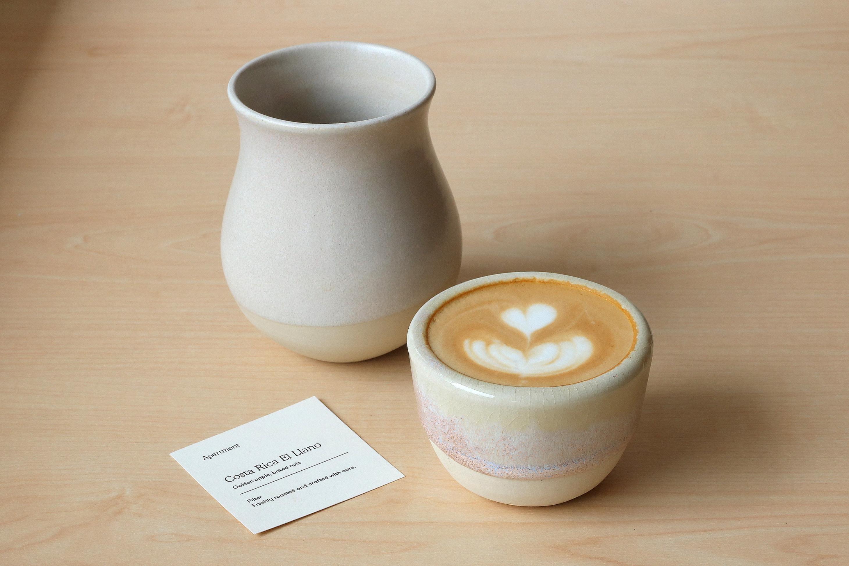 A latte featuring heart-shaped foam art sits in a textured ceramic cup next to a matching pitcher on a light wooden surface. A small white card identifies the coffee as "Costa Rica El Llano" and notes flavor profiles of golden apple and baked nuts.