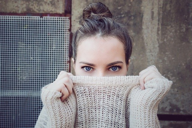 A woman with blue eyes, partially conceals her face with a cotton knitwear sweater