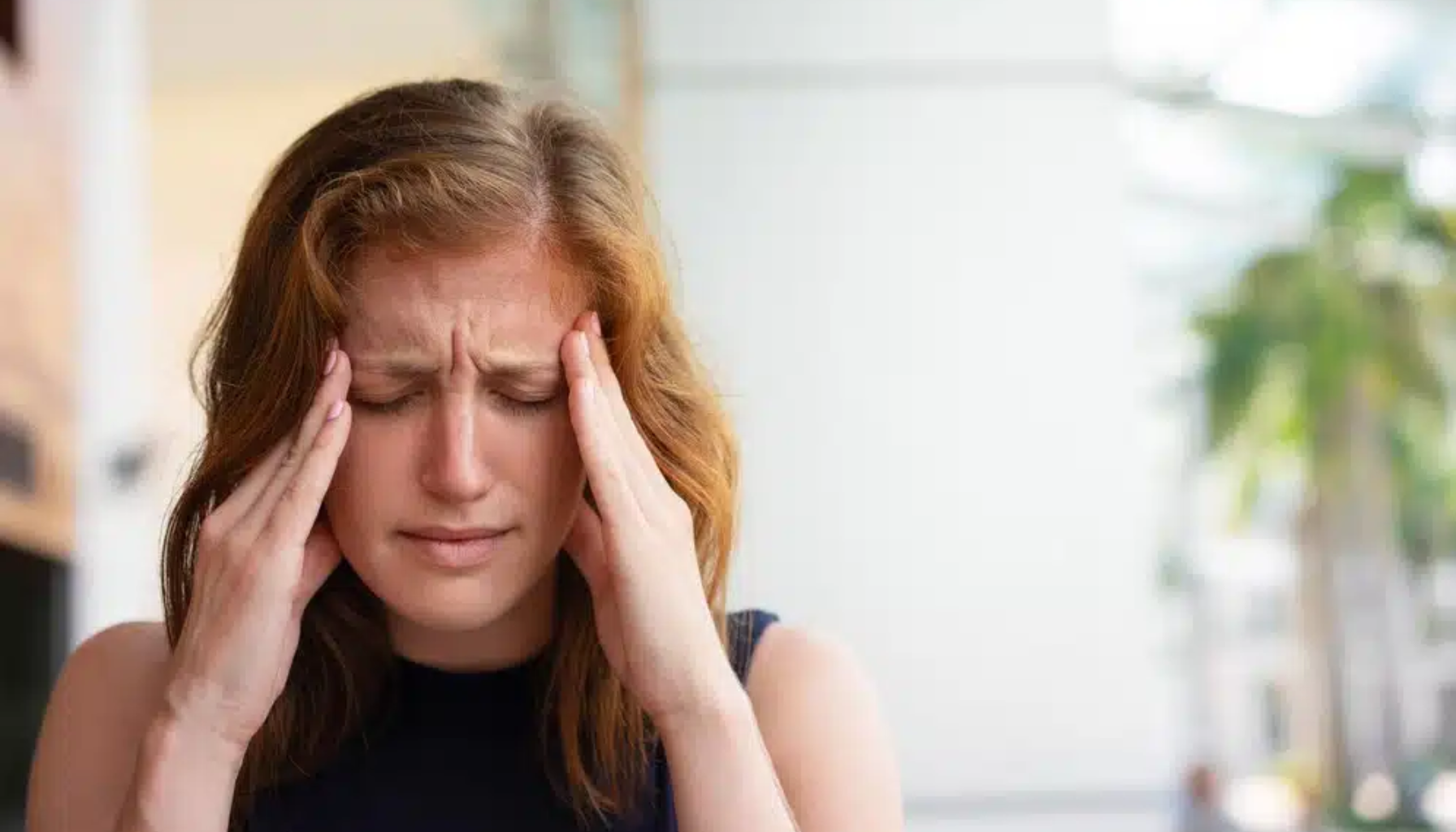 Kvinne med rødt hår holder hendene mot hodet og lukker øynene, tydelig plaget av sterk hodepine eller migrene.