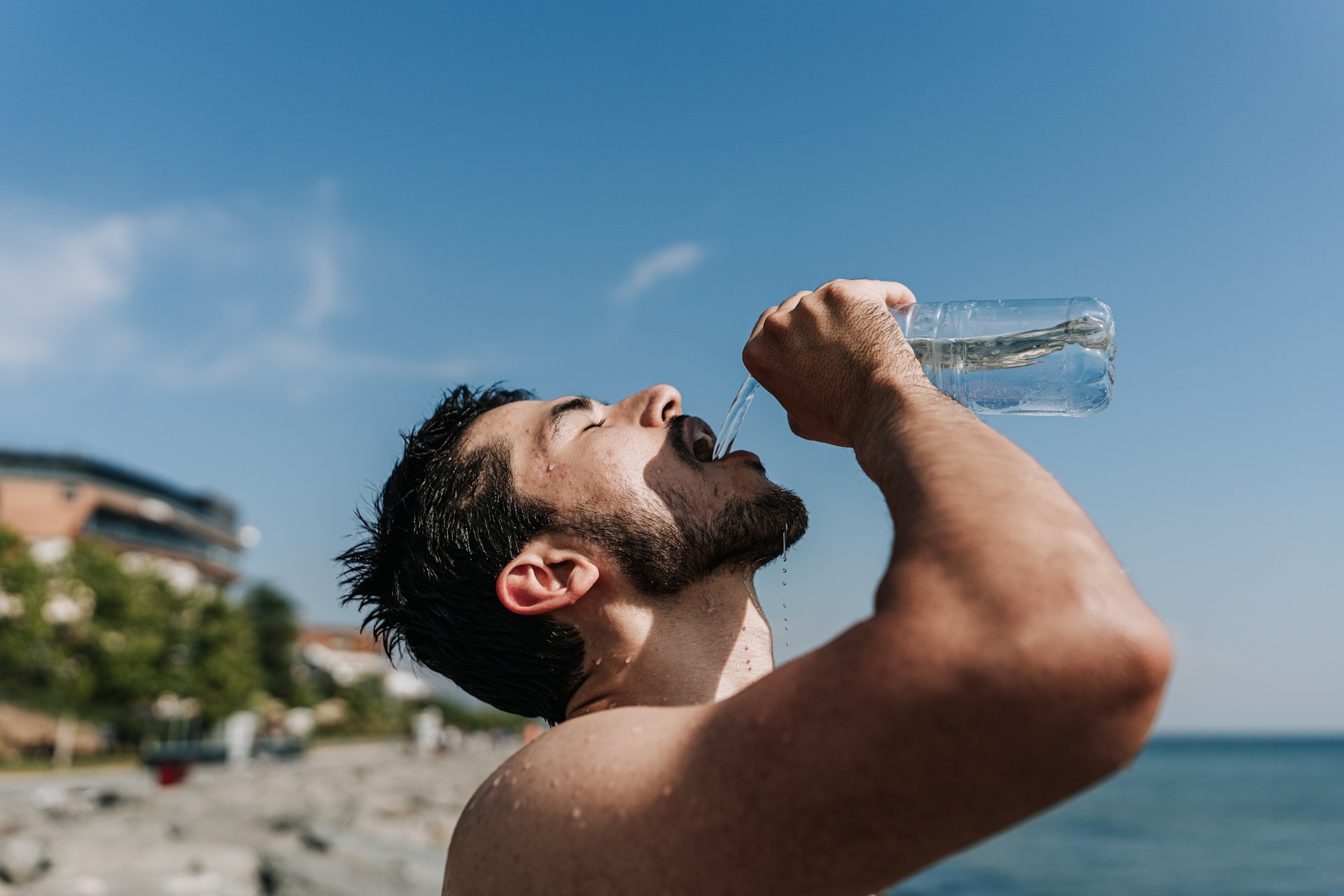 A man pouring a bottle of water into his mouth during a hot day at the beach - https://unsplash.com/photos/a-man-drinking-water-from-a-bottle-on-the-beach-UhXmQeJ6pc0