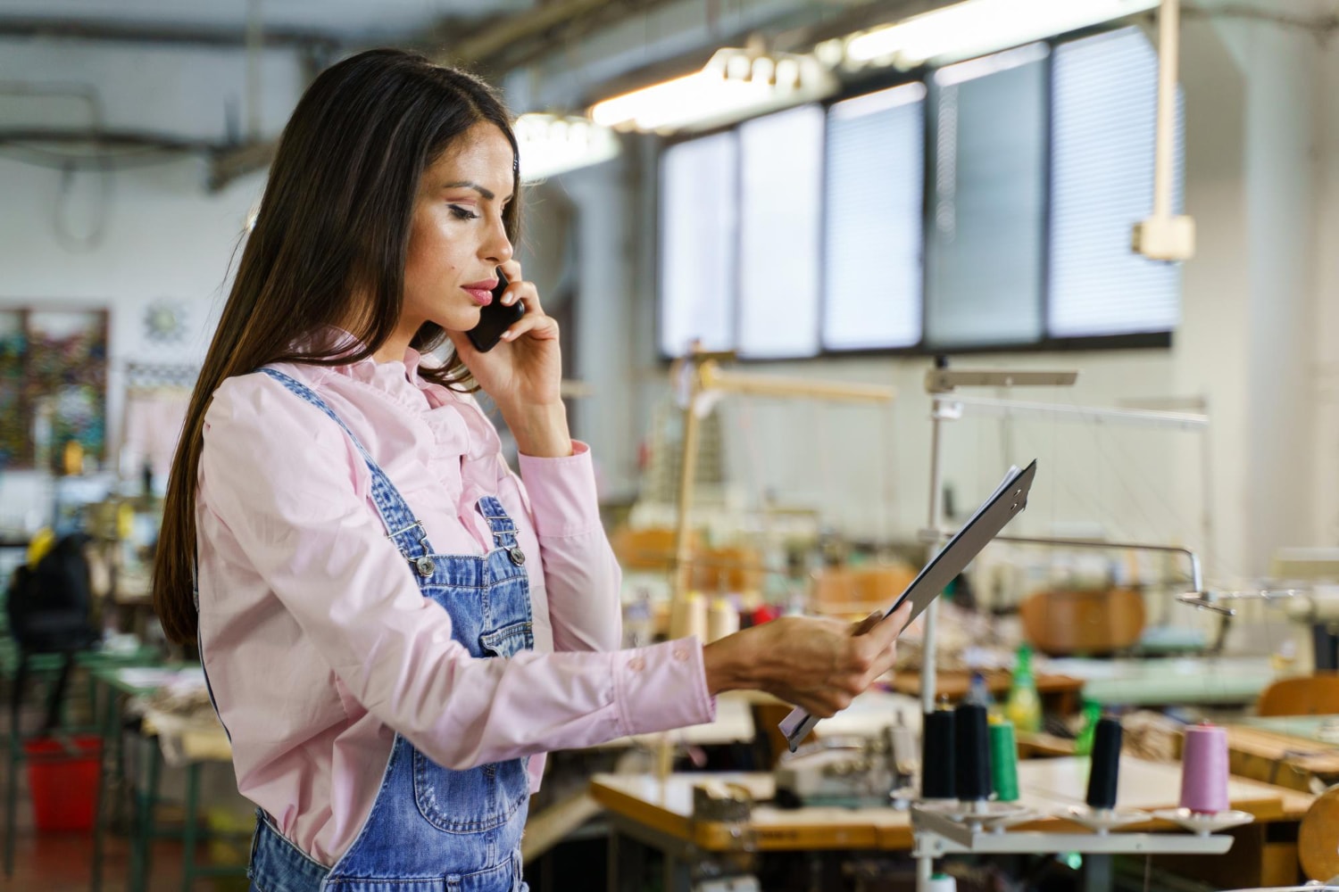 A woman in denim overalls and a pink blouse speaks on her mobile phone while reviewing a clipboard in a sewing factory. Cloud-based communication through UCaaS allows her to manage orders and production workflows efficiently, demonstrating the unified communications benefits of mobile cloud telephony.